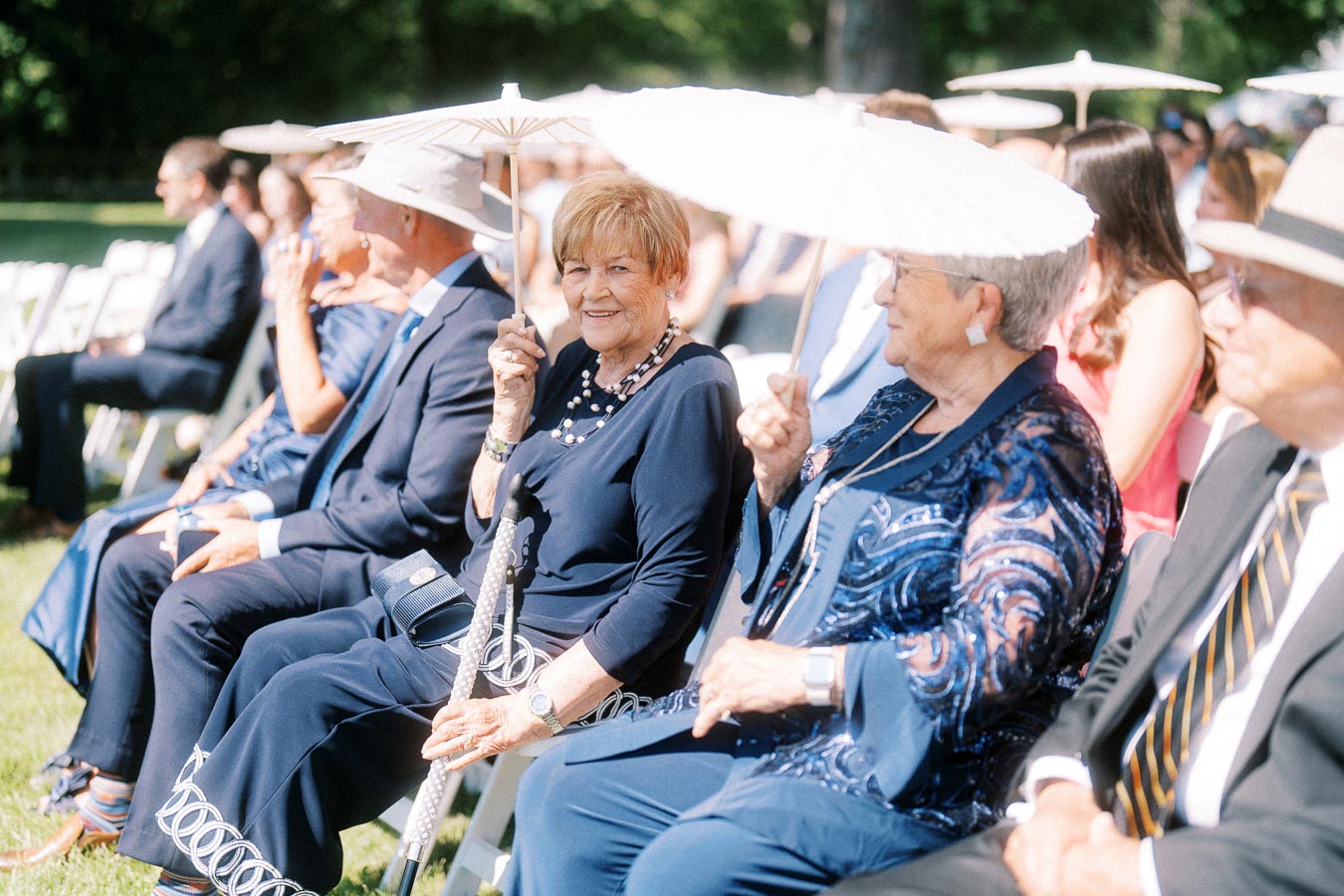 Wedding guests seated outdoors on a lawn in Chatham-Kent, Ontario, holding white parasols during a sunny private estate ceremony.