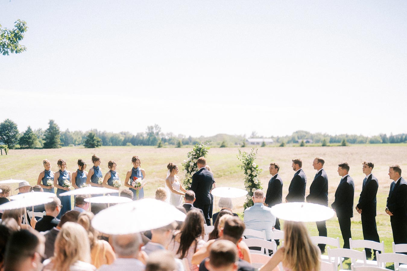 Outdoor wedding ceremony on a private estate near Chatham-Kent, Ontario, with bride, groom, wedding party, guests, white chairs, and floral arches in a sunny farm field.