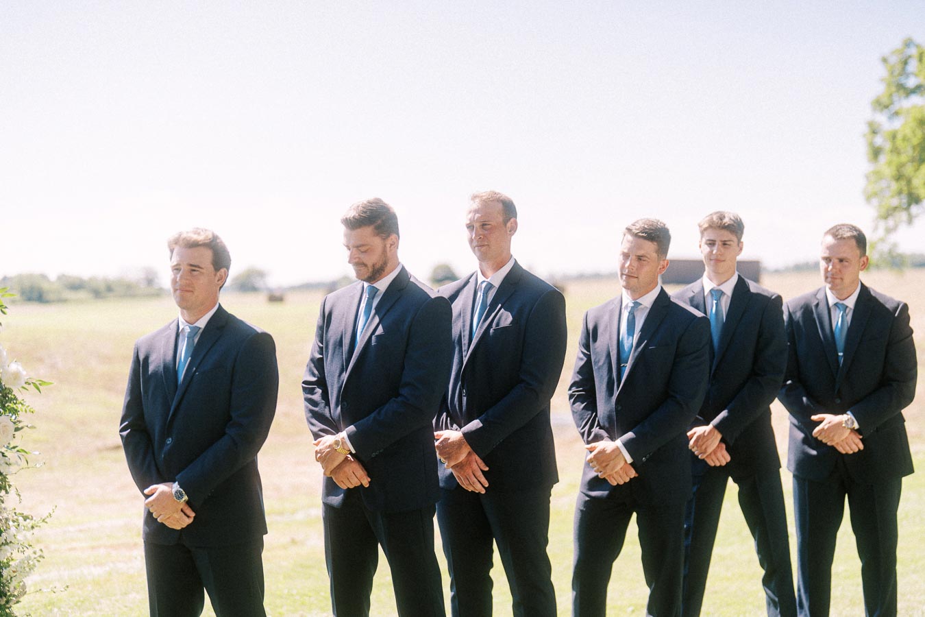 Six groomsmen in dark suits with blue ties stand outdoors in a rural field during a wedding ceremony near Chatham-Kent, Ontario, Canada.