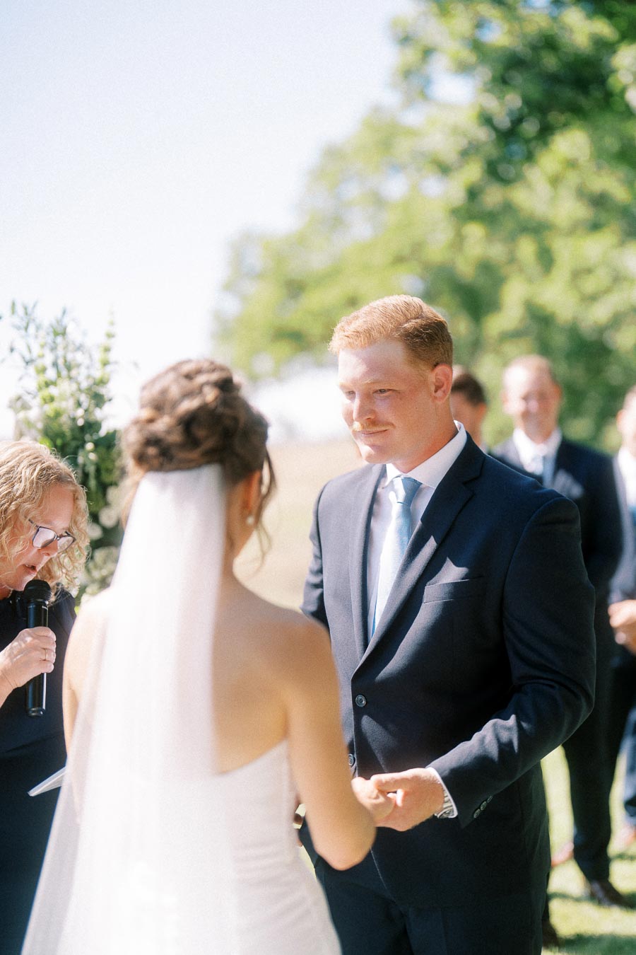 Bride and groom holding hands during an outdoor wedding ceremony near Chatham-Kent, Ontario, with officiant and guests in a sunny garden setting.