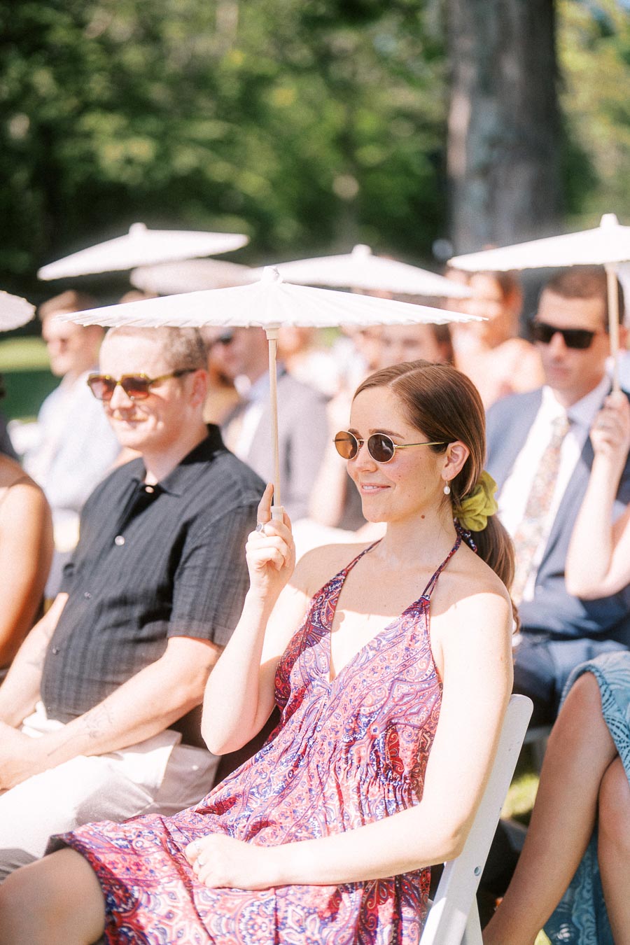 Wedding guests sitting outdoors holding white parasols during a sunny private estate ceremony near Chatham-Kent, Ontario, Canada.