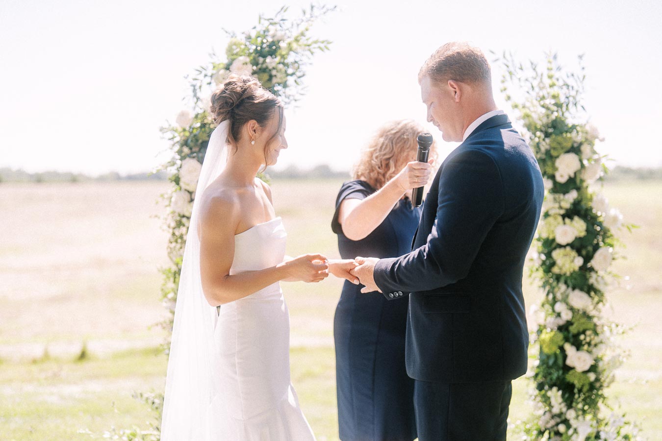 Bride and groom exchanging rings during an outdoor wedding ceremony near Chatham-Kent, Ontario, with officiant and floral arch.