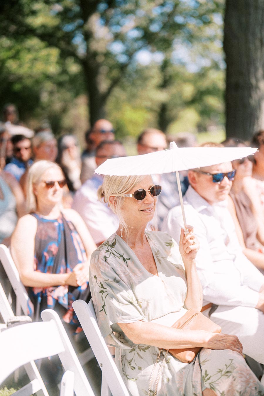 Wedding guests seated outdoors at a private estate ceremony near Chatham-Kent, Ontario, with a woman holding a white parasol in sunlight.