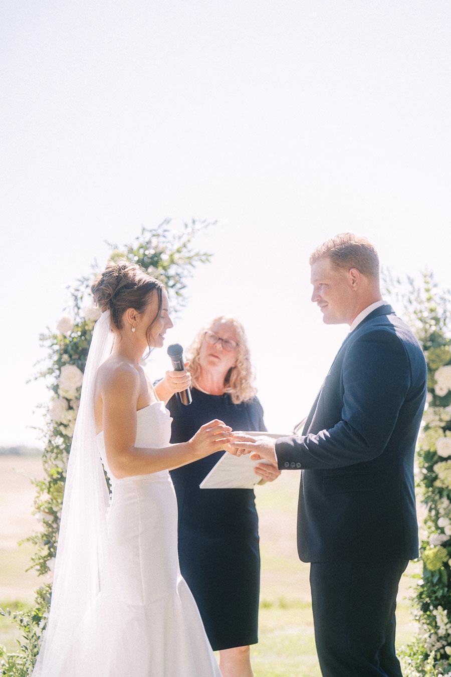Bride and groom exchange rings during an outdoor wedding ceremony in Chatham-Kent, Ontario, with an officiant and floral arch.