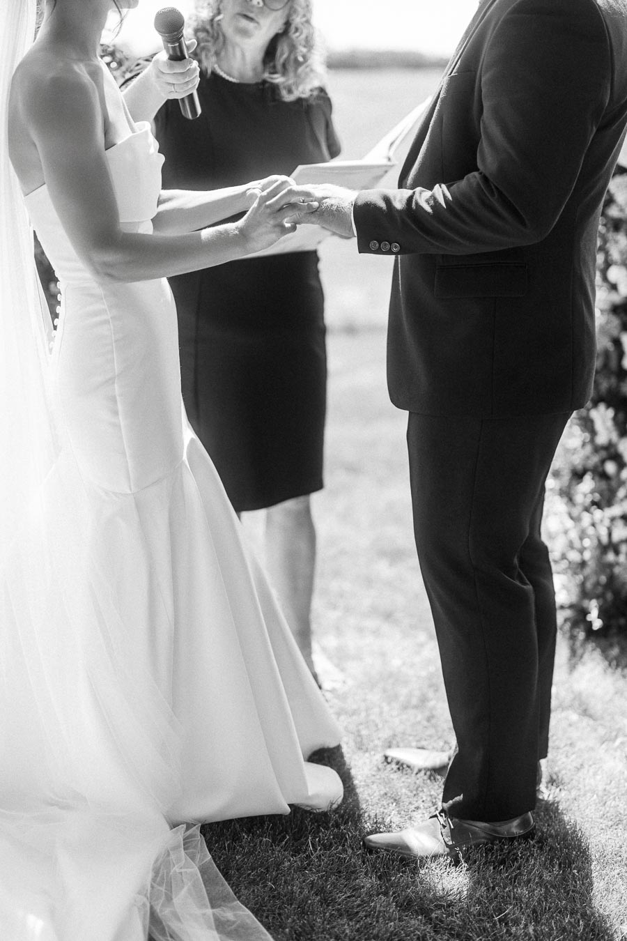 Black and white outdoor wedding ceremony with bride and groom holding hands while an officiant holds a microphone on a grassy private property near Chatham-Kent, Ontario.