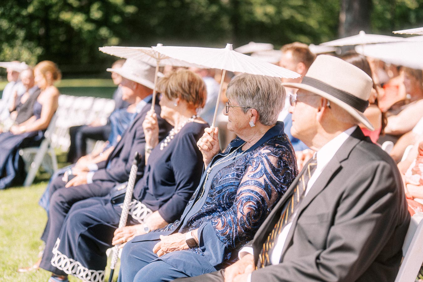 Wedding guests seated outdoors on a sunny lawn holding white parasols during a private estate ceremony near Chatham-Kent, Ontario, Canada.