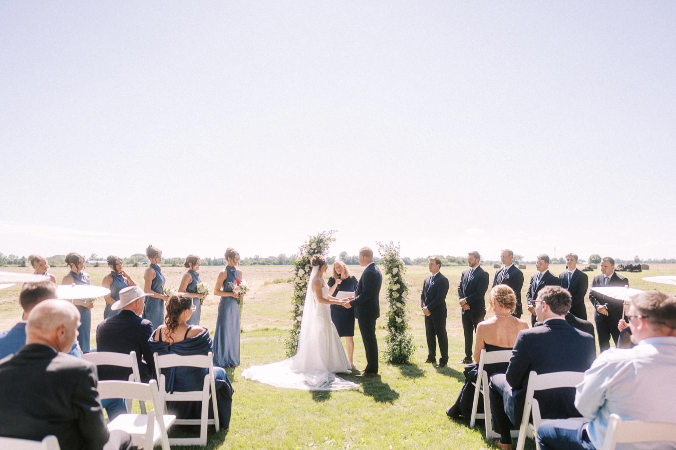 Outdoor wedding ceremony on a private rural estate near Chatham-Kent, Ontario, with bride and groom exchanging vows under a floral arch before seated guests.