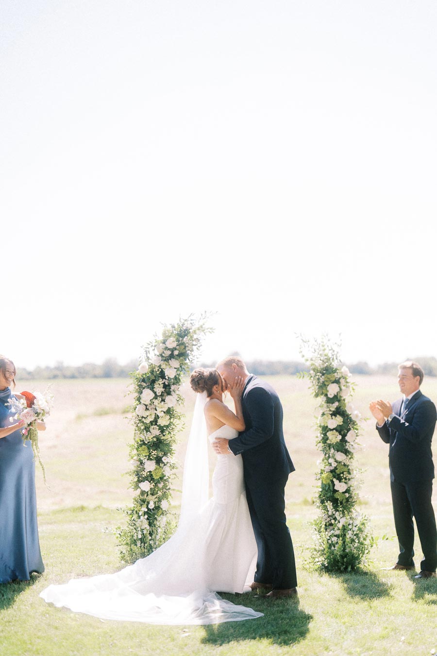 Bride and groom kiss during an outdoor wedding ceremony on a private estate near Chatham-Kent, Ontario, with floral pillars and attendants.