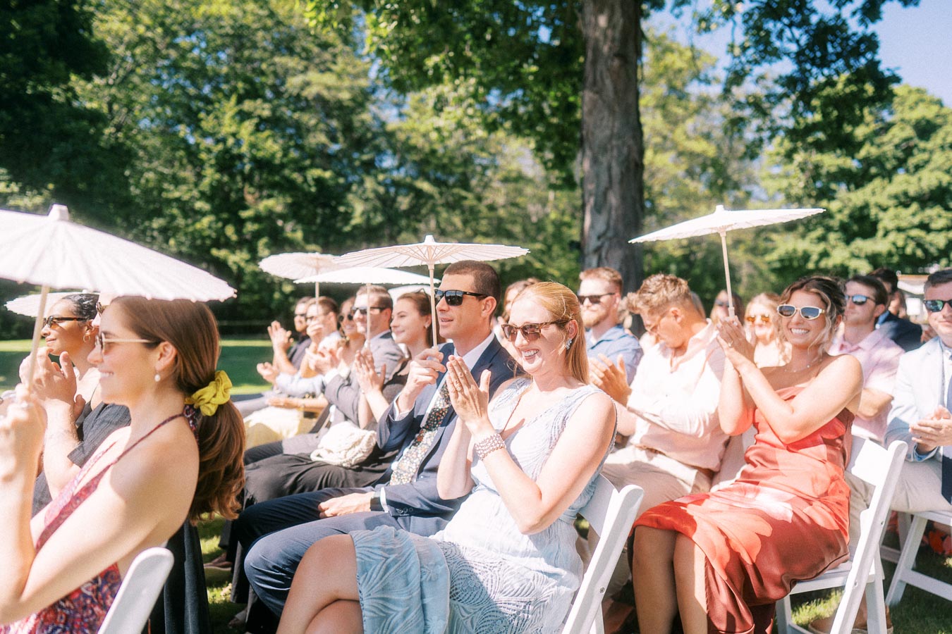 Wedding guests seated on white chairs applaud during an outdoor ceremony on a sunny private estate near Chatham-Kent, Ontario, Canada.