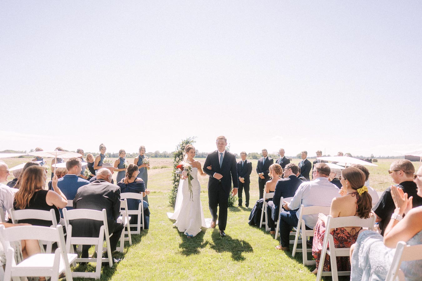 Bride and groom walking down the aisle in an outdoor wedding ceremony, with guests seated on white chairs under a clear blue sky, surrounded by greenery and bridesmaids and groomsmen looking on.