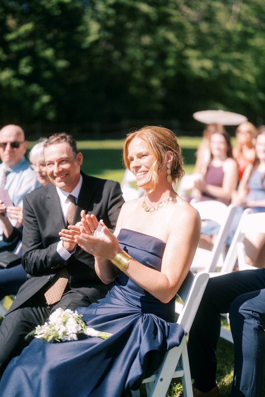 Wedding guests applauding during an outdoor private estate ceremony near Chatham-Kent, Ontario, seated on white chairs in sunny garden setting.