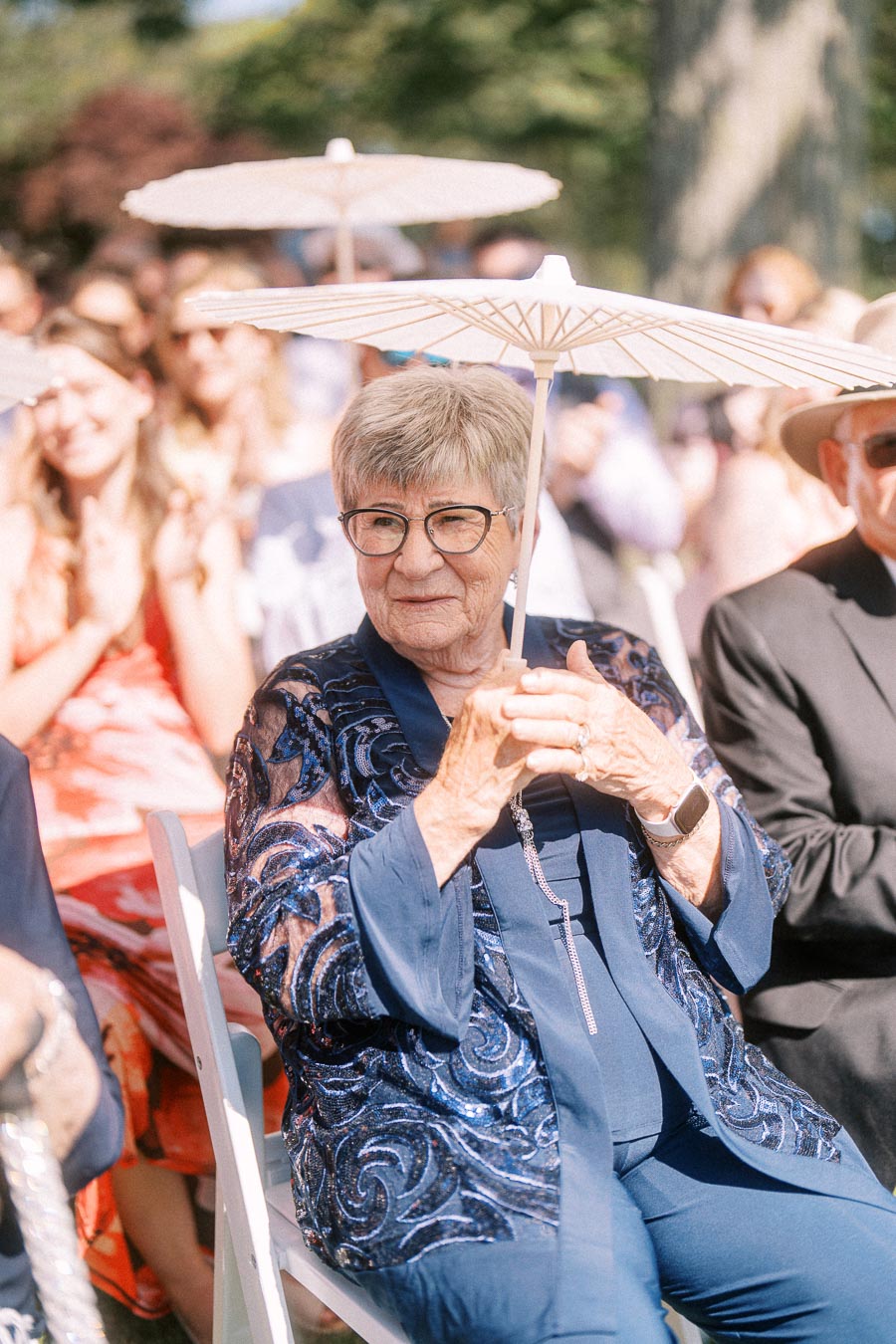 Elderly woman wearing glasses holds a white parasol while seated among guests at an outdoor wedding ceremony in Chatham-Kent, Ontario.