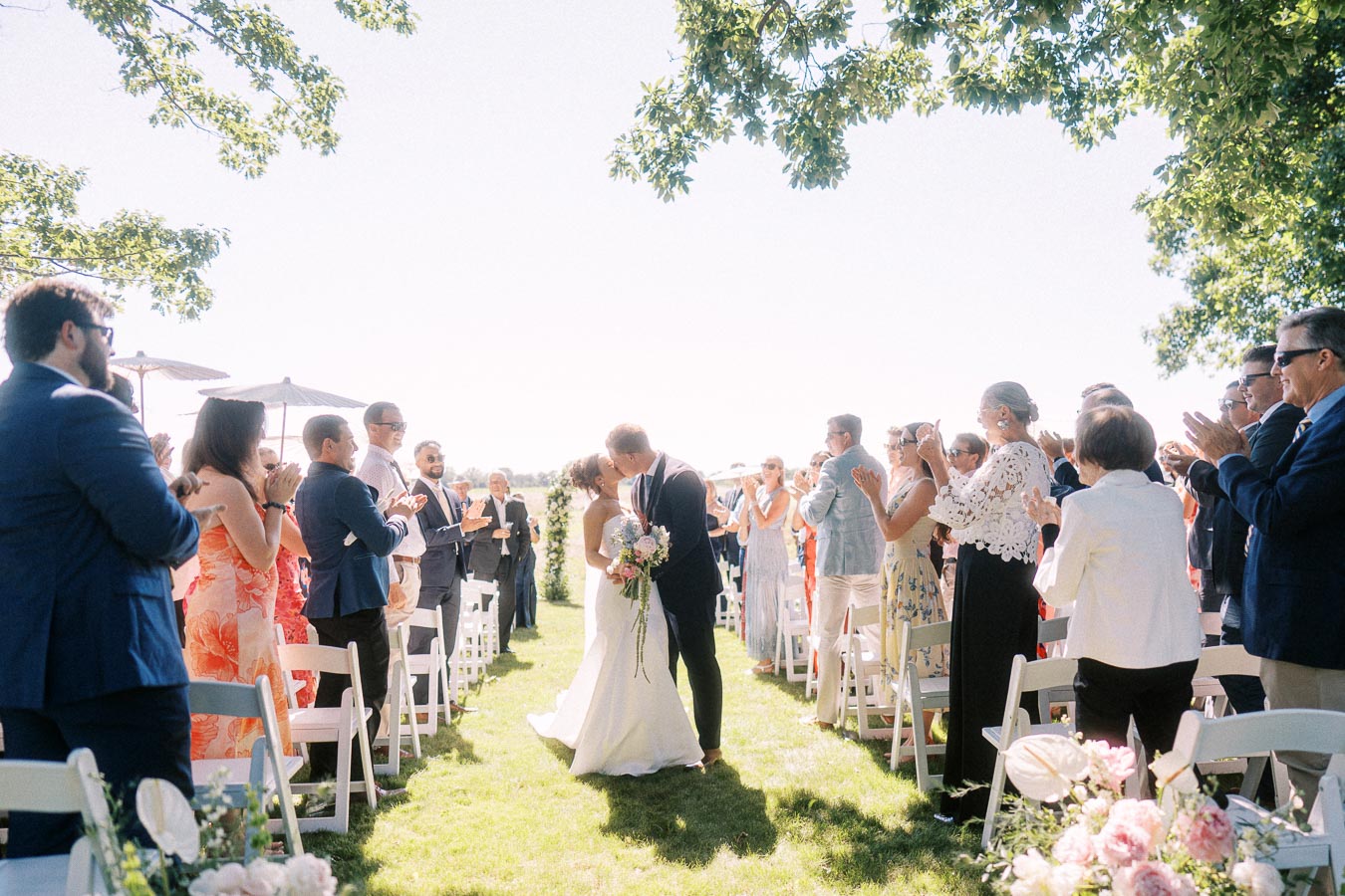 Bride and groom kiss during an outdoor wedding ceremony as guests applaud beside white chairs on a sunny private estate lawn near Chatham-Kent, Ontario.