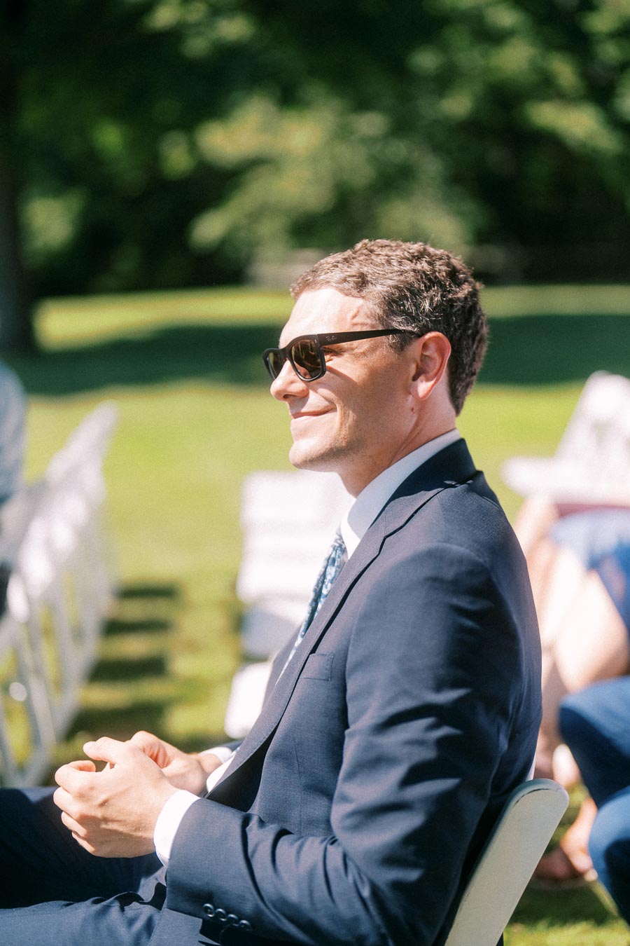 Man in navy suit and sunglasses seated on a white chair at an outdoor wedding ceremony in Chatham-Kent, Ontario, Canada.
