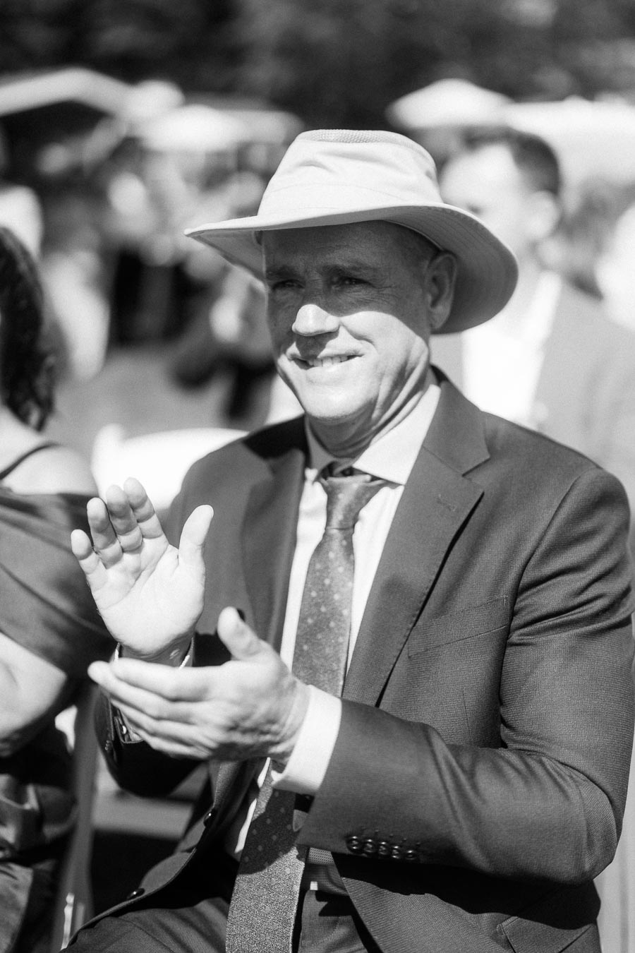 Black and white portrait of a male wedding guest in a suit and hat clapping during an outdoor ceremony near Chatham-Kent, Ontario.