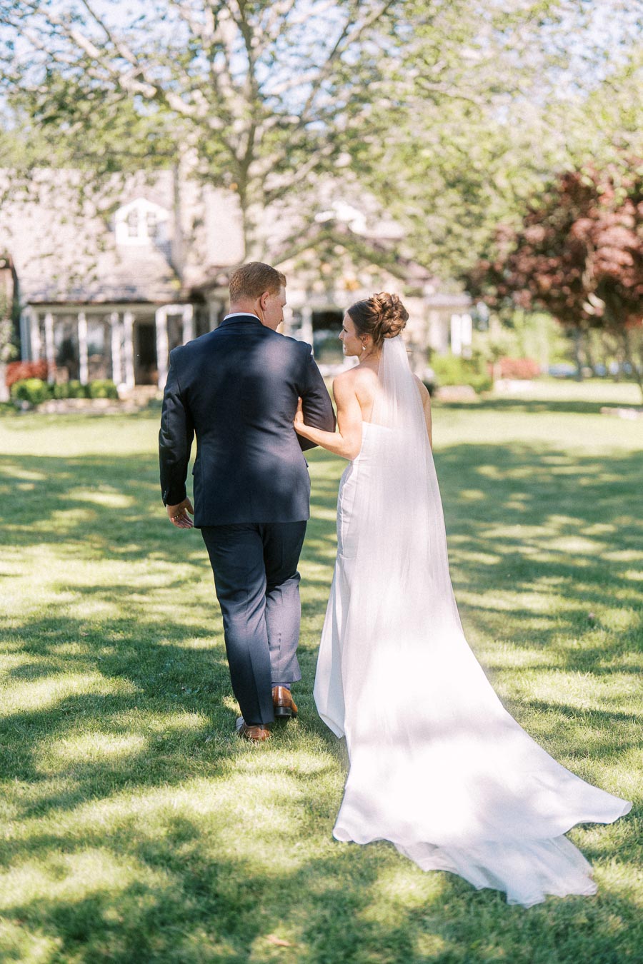 Bride in a white gown and veil walks arm in arm with groom in a navy suit across a sunny lawn at a private estate in Chatham-Kent, Ontario.
