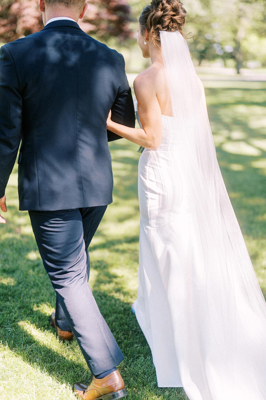 Bride in white gown and veil walks arm in arm with groom in navy suit across a sunny grassy outdoor wedding lawn in Chatham-Kent, Ontario.
