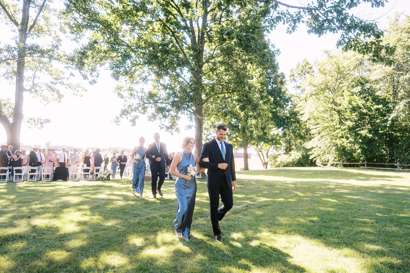 Outdoor wedding procession on a private estate lawn near Chatham-Kent, Ontario, with bridesmaids, groomsmen, guests, trees, and white chairs.