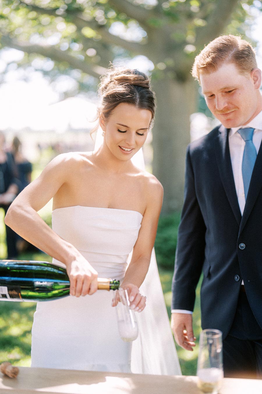 Bride pours sparkling wine into a flute while groom watches during an outdoor private estate wedding near Chatham-Kent, Ontario.