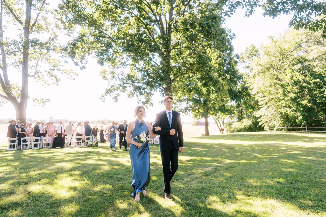 Bridesmaid and groomsman walking arm in arm across a sunny lawn during an outdoor wedding ceremony near Chatham-Kent, Ontario.