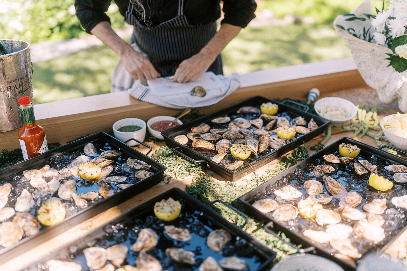 Outdoor wedding oyster bar with chilled oysters, lemons, sauces, flowers, and a server preparing shellfish near Chatham-Kent, Ontario.
