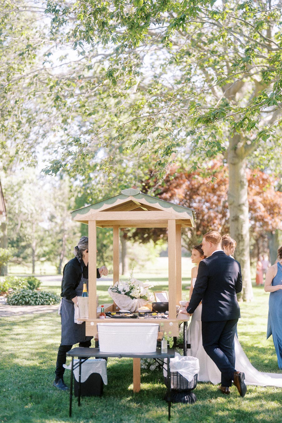 Outdoor oyster bar at a private estate wedding in Chatham-Kent, Ontario, with guests gathered under trees on a sunny lawn.