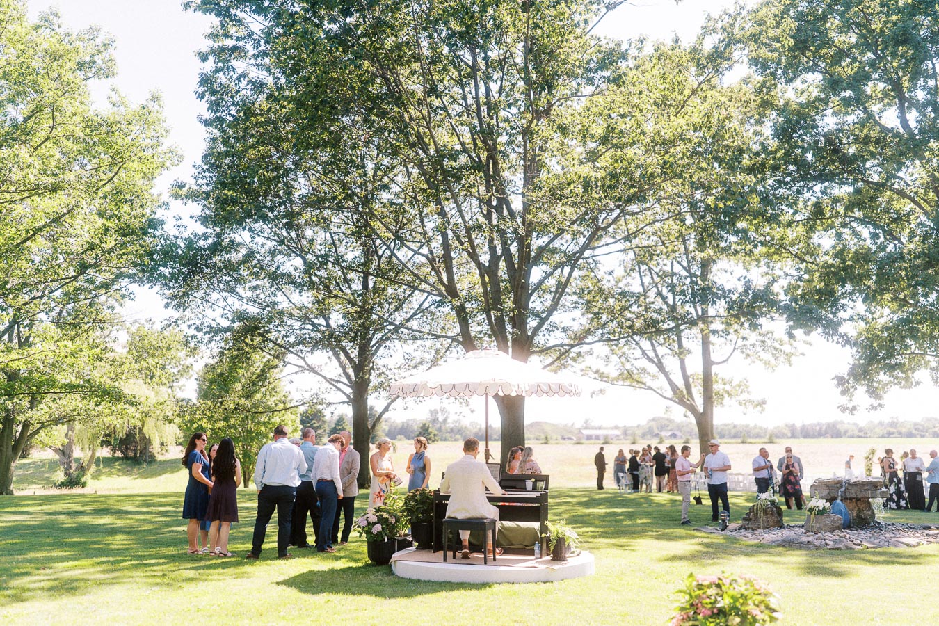 Guests gather on a grassy private estate lawn near Chatham-Kent, Ontario, with a pianist performing under large trees during an outdoor farm wedding.