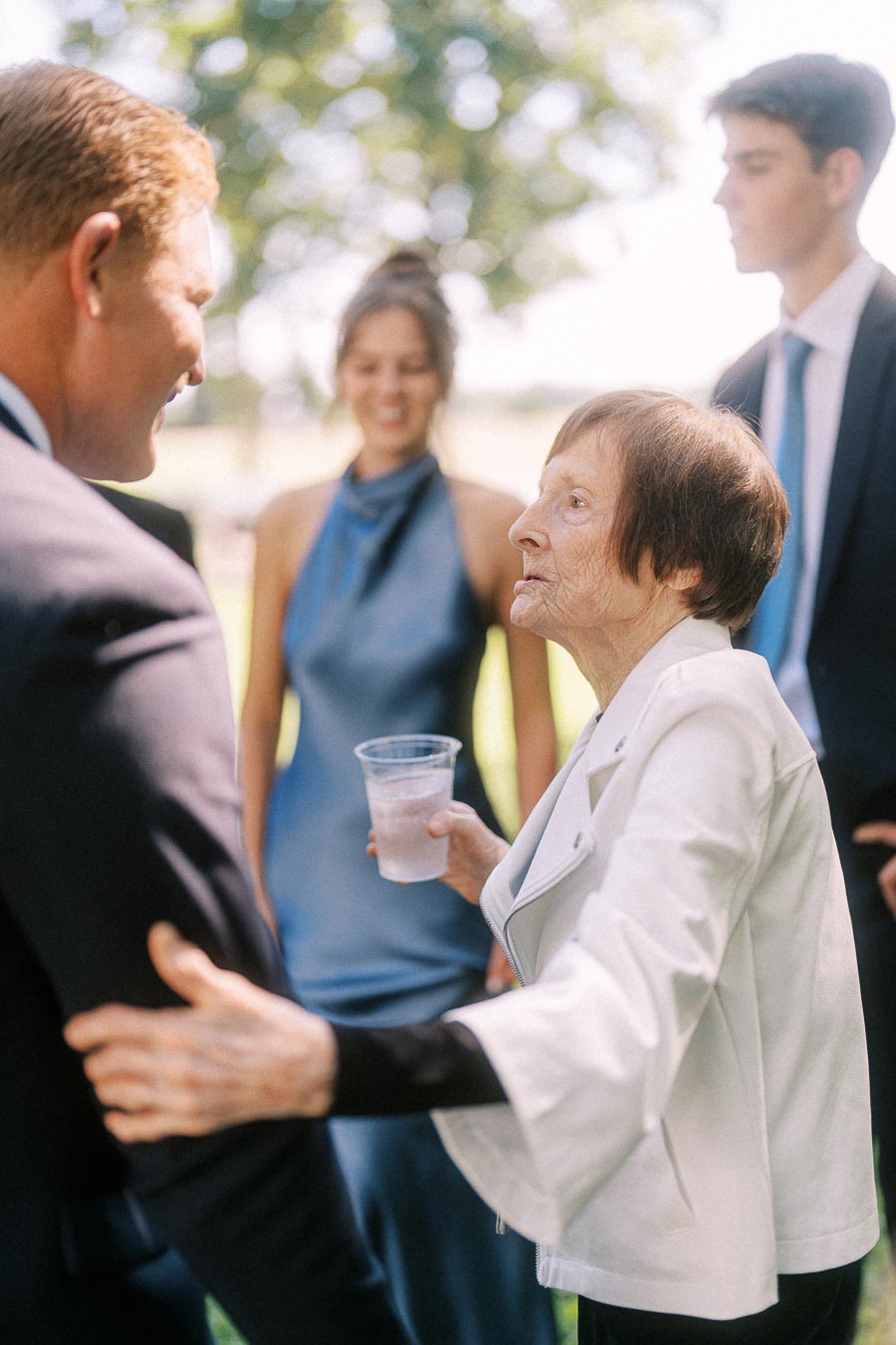 Elderly woman in a white jacket greeting a man in a suit while holding a plastic cup at an outdoor wedding gathering near Chatham-Kent, Ontario.