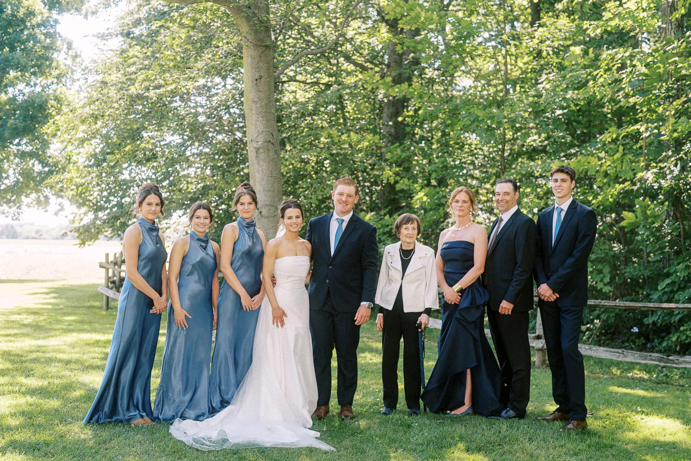 Outdoor wedding party portrait on a private estate lawn near Chatham-Kent, Ontario, with bride, groom, family, and bridesmaids under trees.