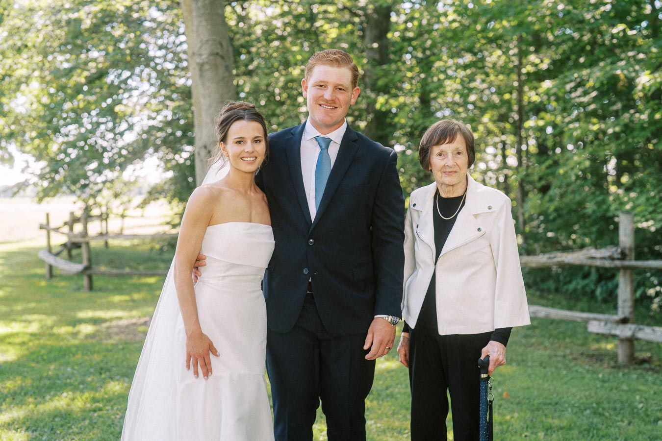 Bride, groom, and elderly woman posing outdoors on a private farm estate near Chatham-Kent, Ontario, during a sunny wedding portrait session.