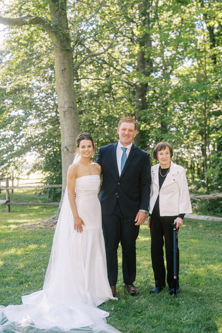 Bride, groom, and elderly woman standing together on grass beneath trees at an outdoor private estate wedding near Chatham-Kent, Ontario.
