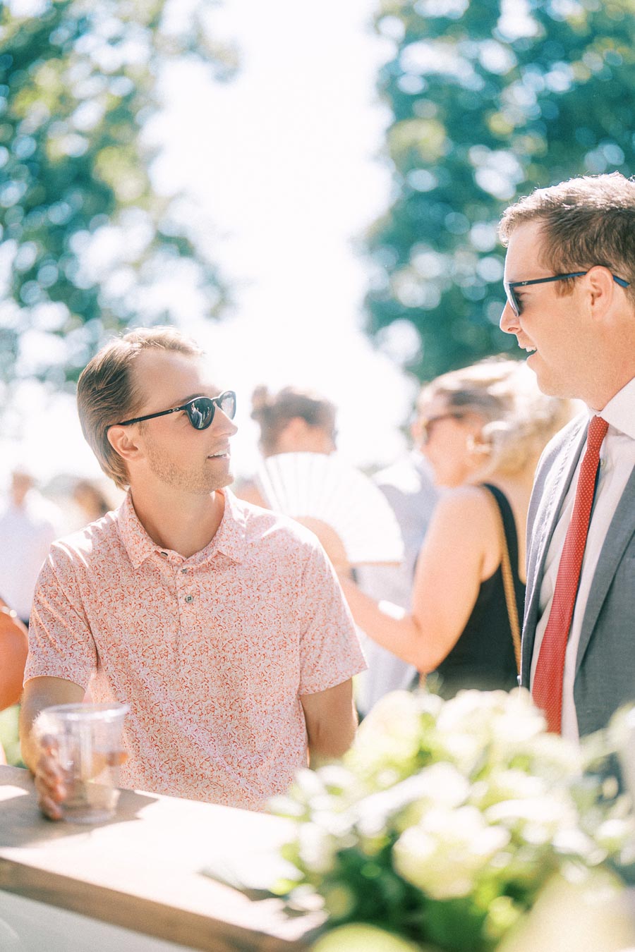Two men in sunglasses talk at an outdoor wedding reception near Chatham-Kent, Ontario, with guests and flowers in bright sunlight.