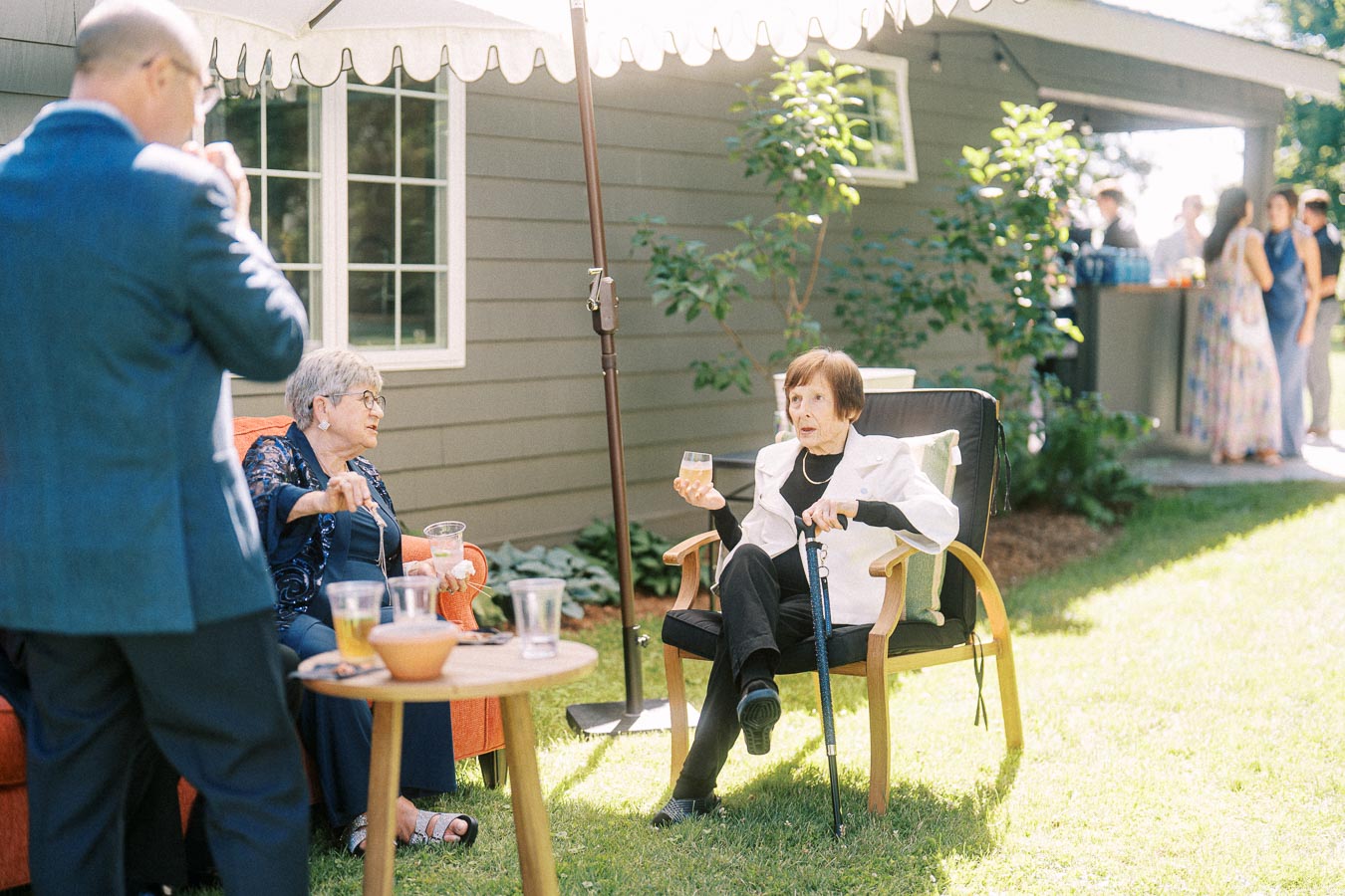 Wedding guests relaxing outdoors on a private property near Chatham-Kent, Ontario, with elderly women seated under a patio umbrella beside a house.