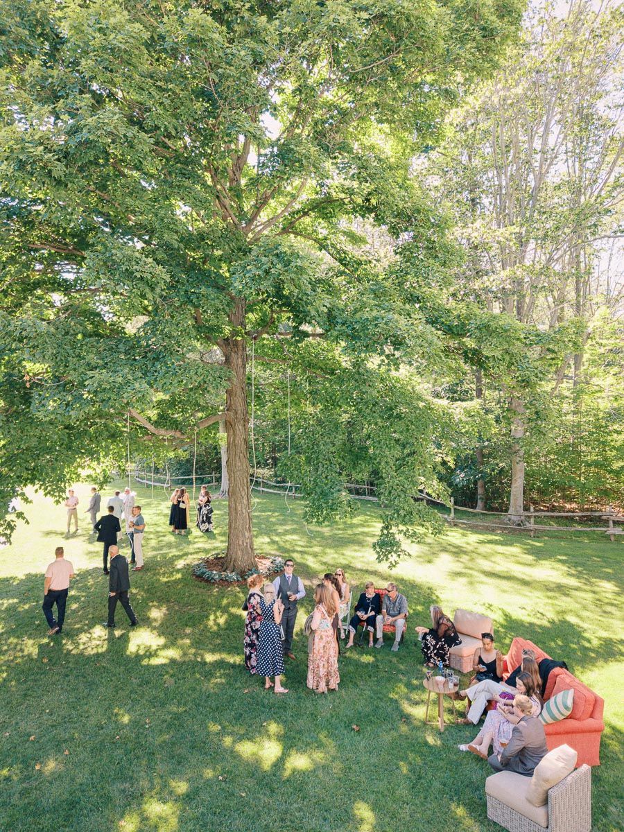Wedding guests mingle on a shaded lawn beneath large trees at a private estate outdoor farm wedding near Chatham-Kent, Ontario, Canada.