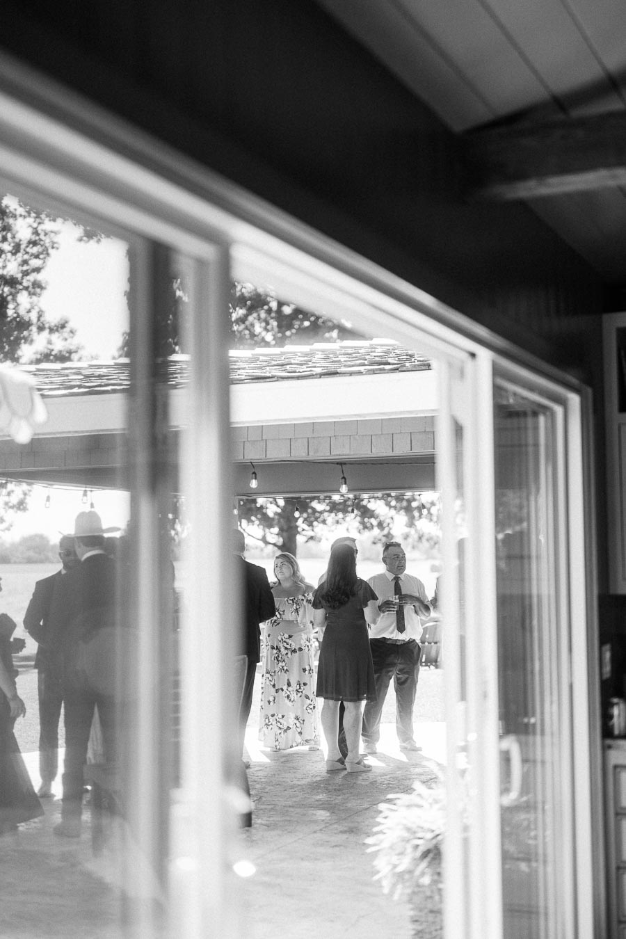 Black and white view through glass doors of wedding guests socializing on a covered patio at a private estate near Chatham-Kent, Ontario.