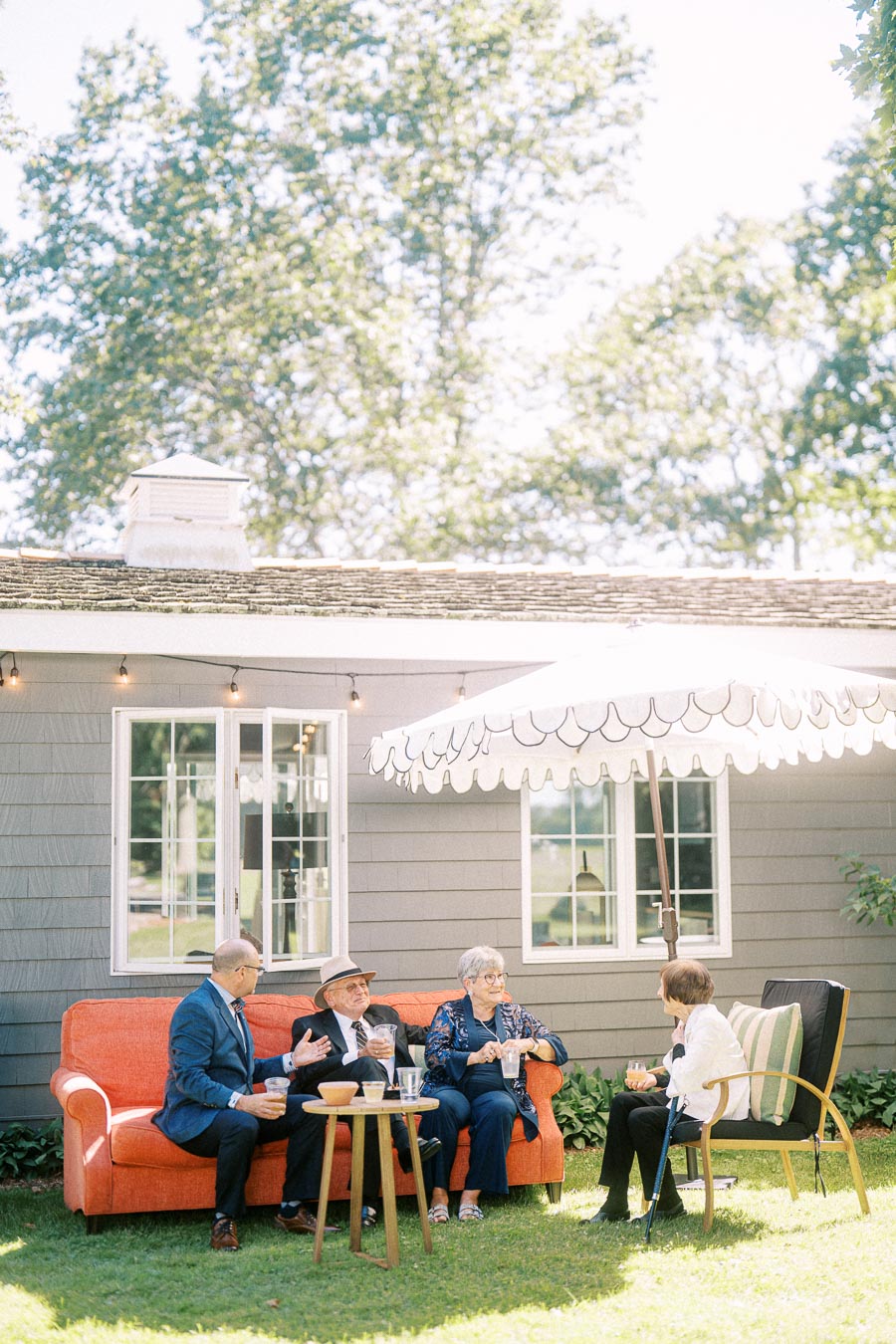 Wedding guests relaxing outdoors on a private estate lawn near Chatham-Kent, Ontario, beside a house with sofa, chairs, umbrella, and string lights.