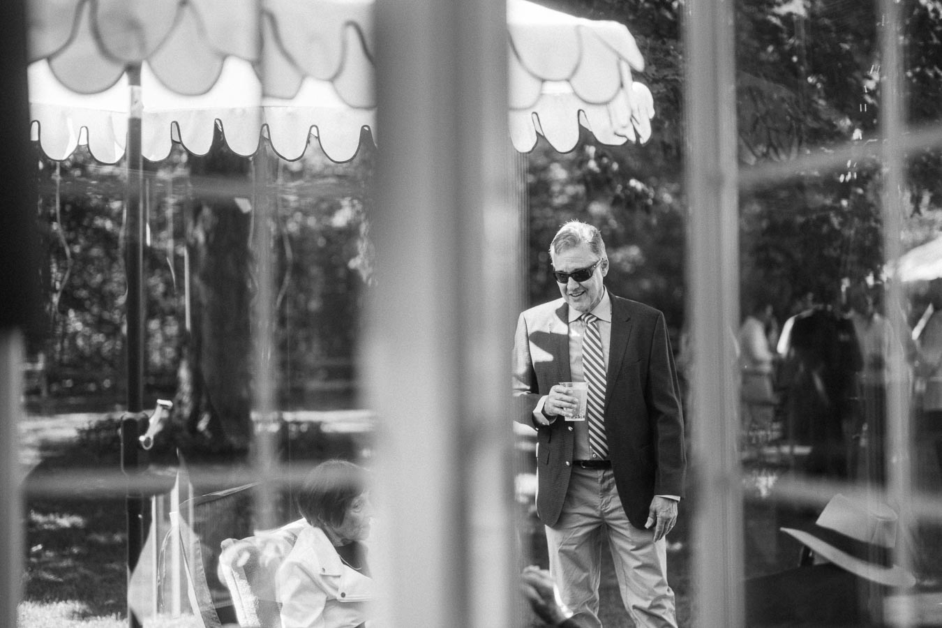 Black and white candid view through windows of a man in sunglasses holding a drink at an outdoor tented wedding on a private estate near Chatham-Kent, Ontario.