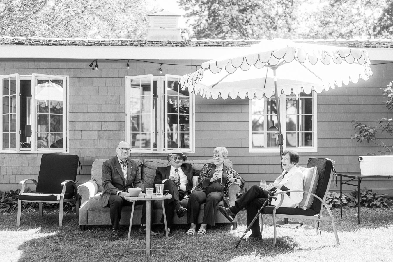 Black and white photo of four senior wedding guests seated on outdoor furniture under a patio umbrella beside a house in Chatham-Kent, Ontario.