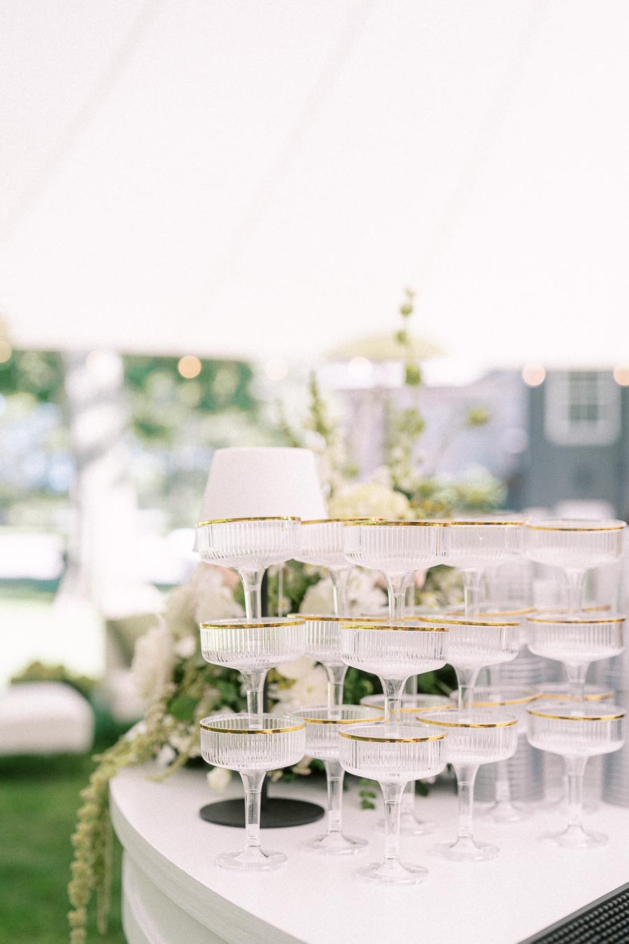 Stacked champagne coupe glasses with gold rims on a white bar inside a tented outdoor wedding reception near Chatham-Kent, Ontario.