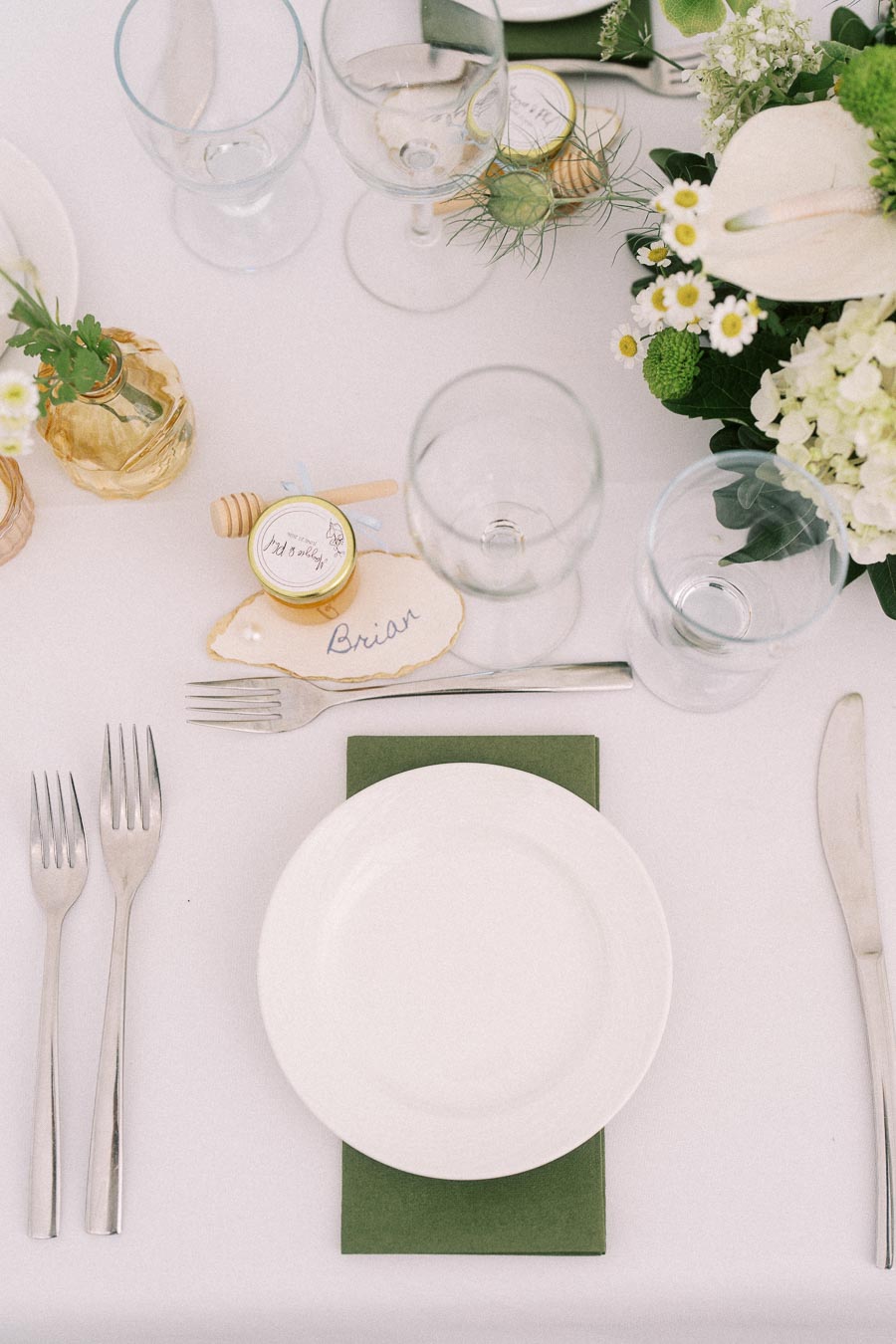 Elegant wedding table setting with white plate, green napkin, glassware, honey favor, place card, and white floral centerpiece.