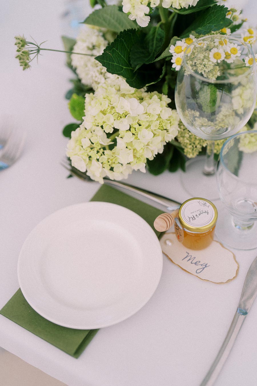 Close-up of an elegant outdoor wedding table setting near Chatham-Kent, Ontario, with white florals, glassware, honey favor, and a handwritten place card.