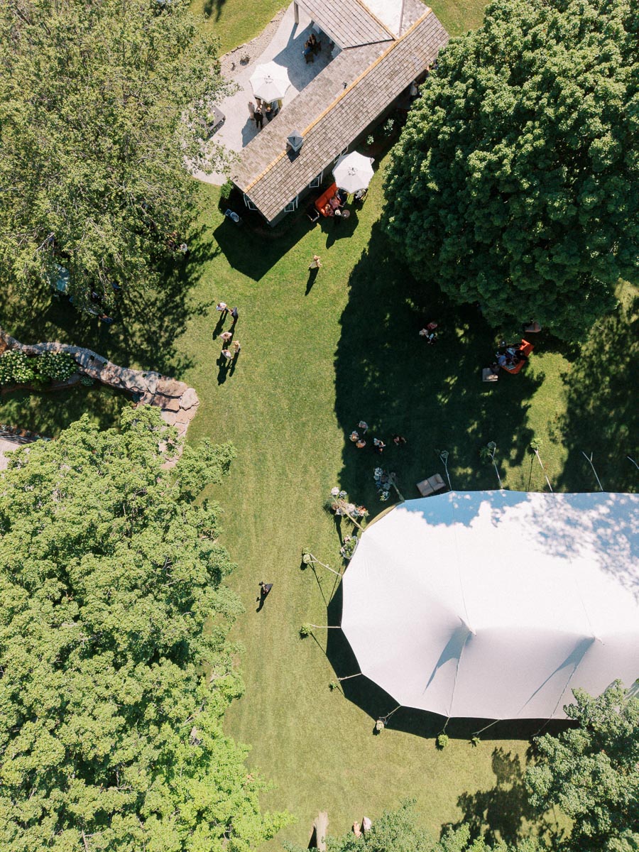 Aerial view of a private estate outdoor wedding setup with a white tent, lawn, trees, guests, and a rustic farm building near Chatham-Kent, Ontario.