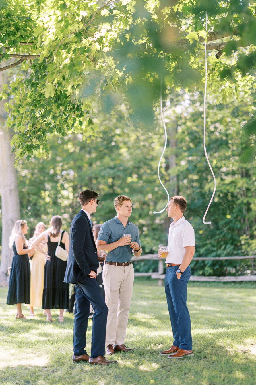 Wedding guests chatting with drinks on a shaded lawn beneath trees at a private estate outdoor wedding near Chatham-Kent, Ontario, Canada.