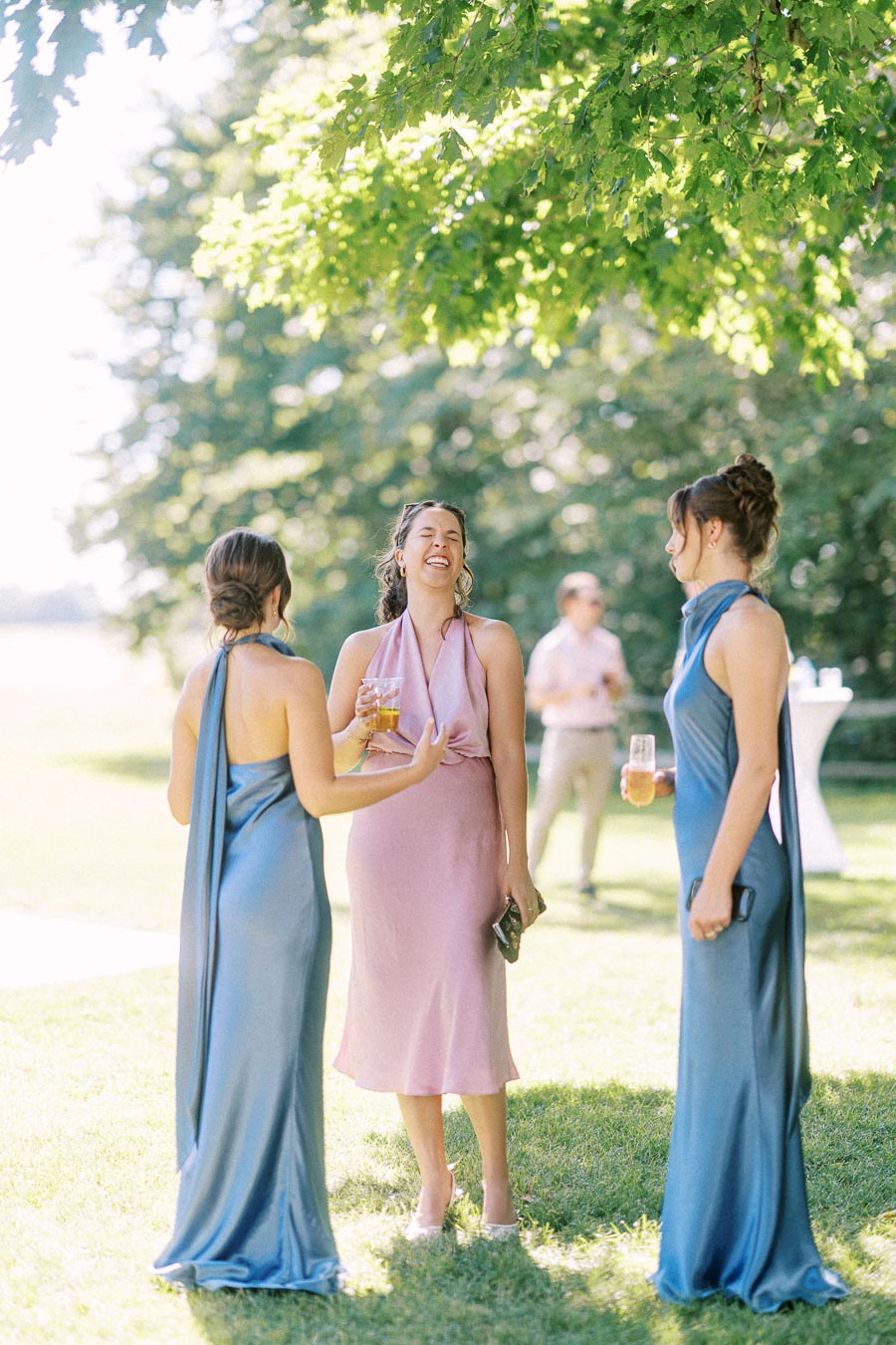 Three women in formal dresses laugh and talk outdoors with drinks during a sunny private estate wedding near Chatham-Kent, Ontario.