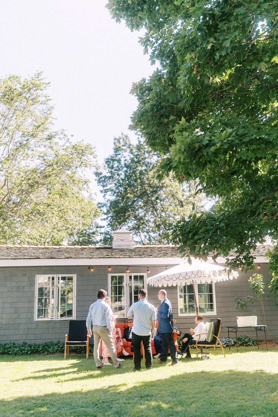 Wedding guests mingle on a sunny lawn beside a private estate house with a patio umbrella and large trees near Chatham-Kent, Ontario.