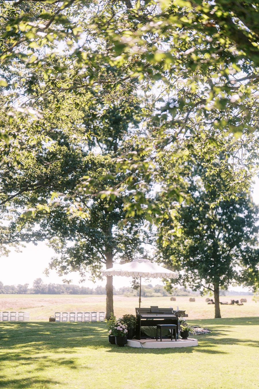 Outdoor farm wedding ceremony setup on a private estate near Chatham-Kent, Ontario, with chairs, piano, umbrella, flowers, trees, and hay bales.