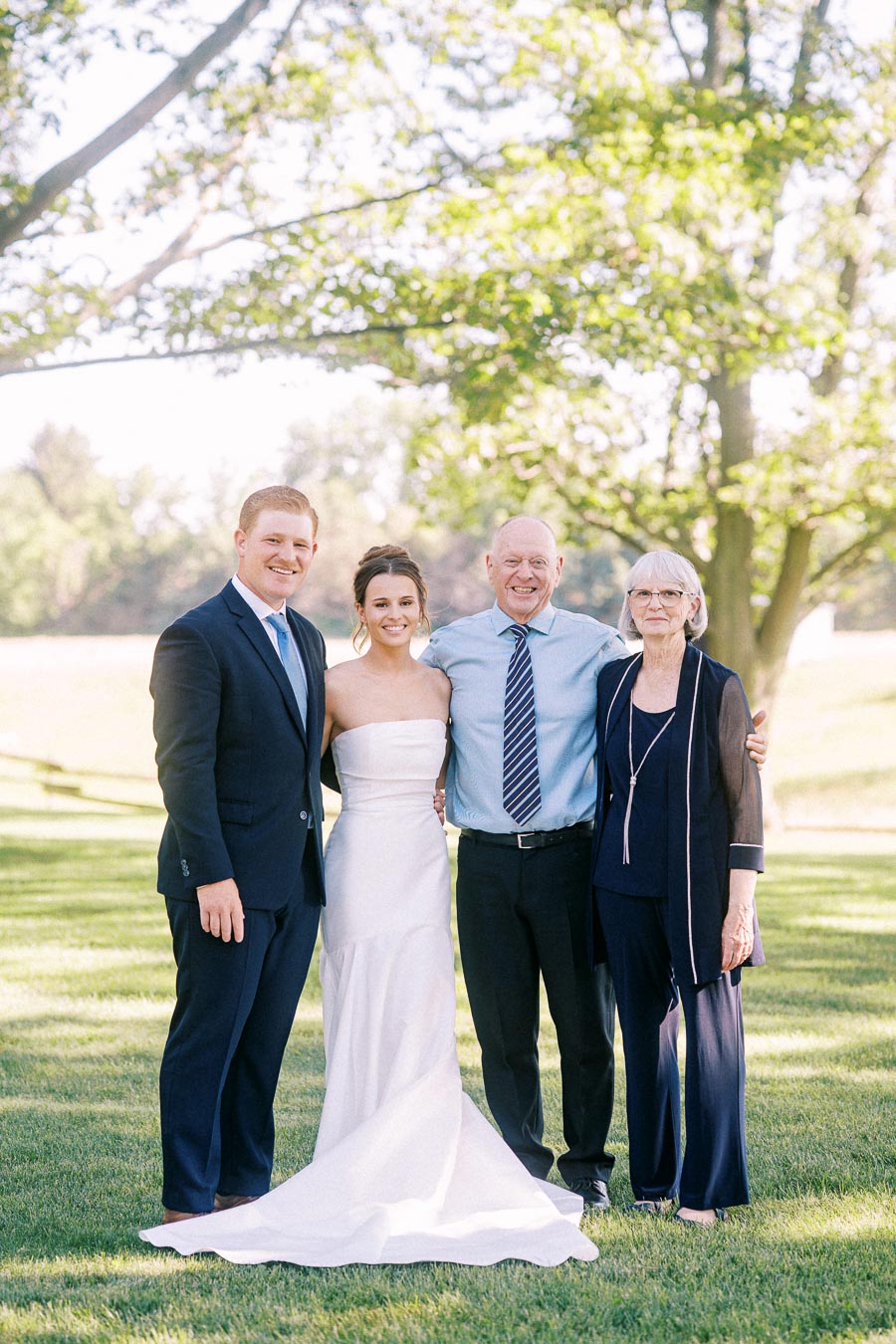 Bride in white strapless wedding dress poses with groom and older couple on a sunny lawn at a private estate near Chatham-Kent, Ontario.