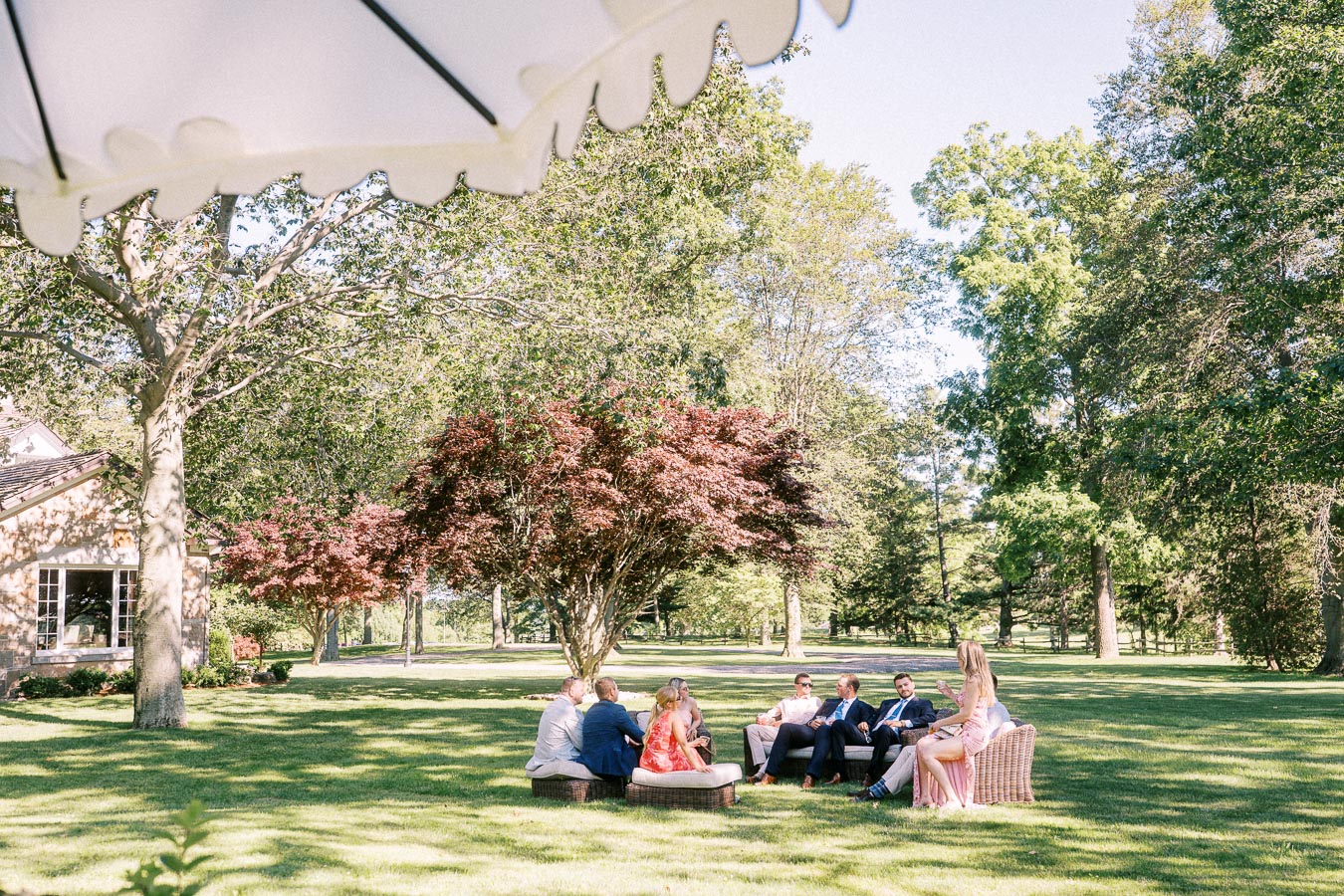 Wedding guests relax on outdoor lounge seating under trees at a private estate near Chatham-Kent, Ontario, with a tent edge above.