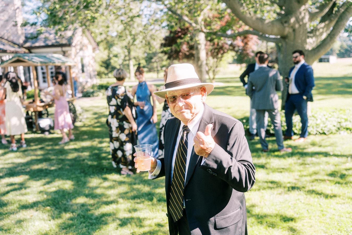 Man in suit and hat holding a drink and gesturing at an outdoor wedding gathering on a private estate lawn near Chatham-Kent, Ontario.