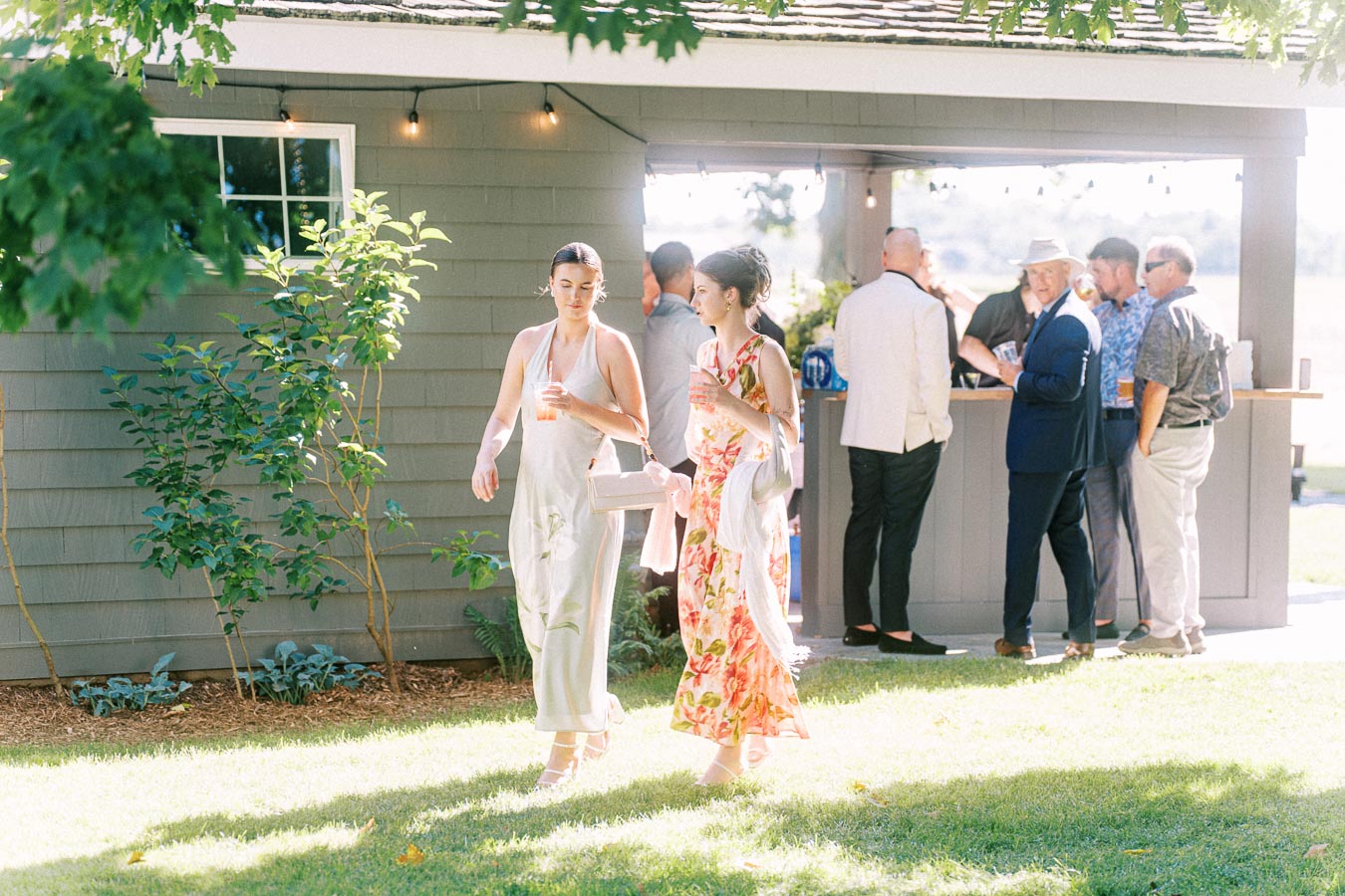Wedding guests mingle outdoors beside a private estate bar near Chatham-Kent, Ontario, with two women walking across a sunny lawn holding drinks.